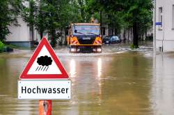 Foto: Hochwasserschild vor überfluteter Straße, Straßenmeisterfahrzeug im Hintergrund